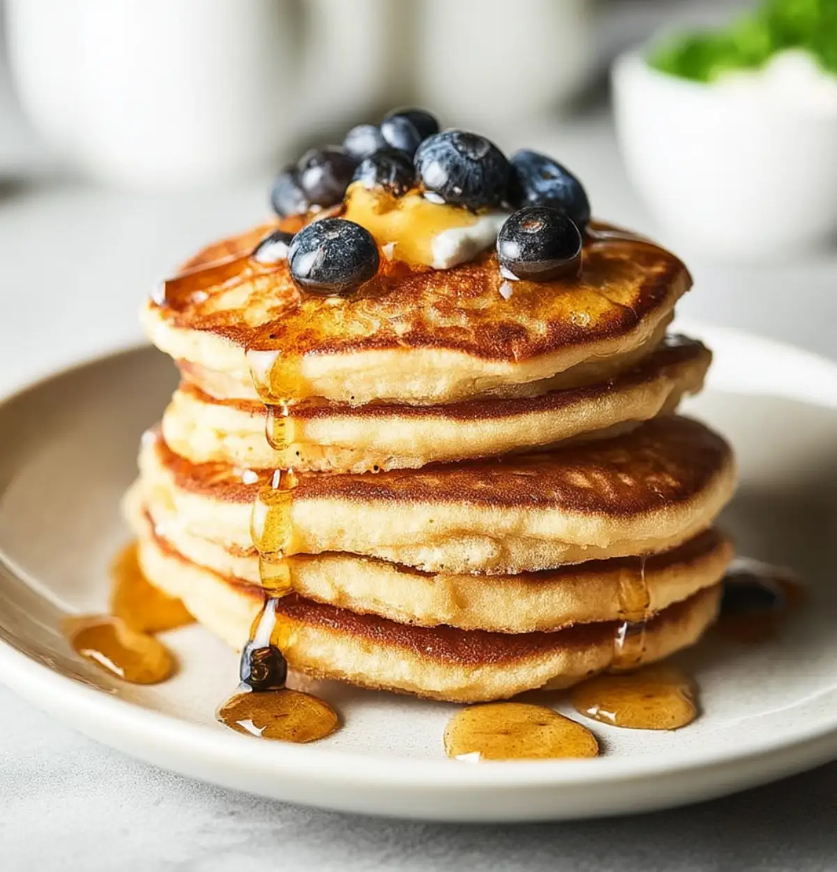 Fluffy stack of high-protein cottage cheese pancakes topped with fresh blueberries and drizzled with golden syrup, served on a white plate.